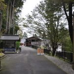 比叡山延暦寺の境内入口の様子。左右には木製の大きな柱が立ち、それぞれに「延暦寺」と「比叡山」と書かれています。奥には参道が続き、木々に囲まれた建物が見えます。道沿いには案内所や休憩所と思われる小さな建物があり、参拝者が歩いている姿も確認できます。周囲は緑豊かで、静かな雰囲気が漂っています。