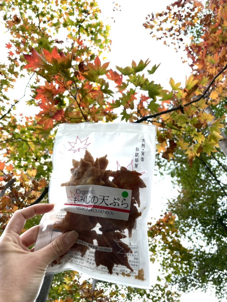 紅葉を背景に「もみじの天ぷら」の入った袋を掲げている。