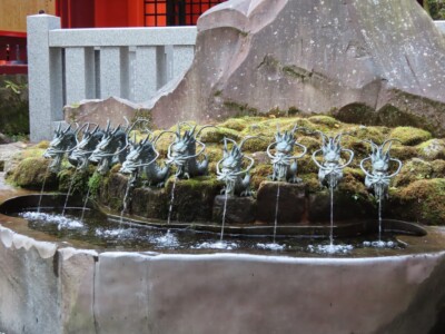 九頭龍神社新宮前にある霊水「龍神水」の手水舎です。苔むした岩から八つの龍の頭が並び、それぞれの口から清らかな水が流れ出しています。背景には神社の建物の一部が見え、神聖な雰囲気が漂っています。