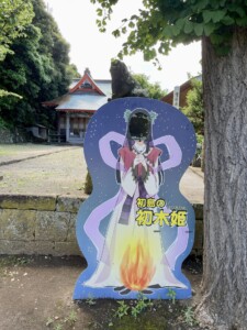 神社の境内に設置された「初島の初木姫」の顔出しパネル。パネルには巫女装束を身に着けたキャラクターが描かれており、背景には夜空と炎が描かれている。顔の部分は穴が開いており、記念写真を撮ることができるようになっている。背後には伝統的な神社の建物と石造りの狛犬が見える。周囲は緑に囲まれており、木陰が心地よい。
