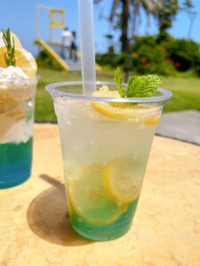 透明なカップに入ったレモンのスライスとミントの葉が浮かぶレモネード。背景には芝生とヤシの木が見え、遠くには観光客がいる。隣にはホイップクリームとレモンのスライスがトッピングされた青いドリンクが少しだけ見える。リフレッシュできるような爽やかな飲み物が、美しい景色とともに楽しめる。