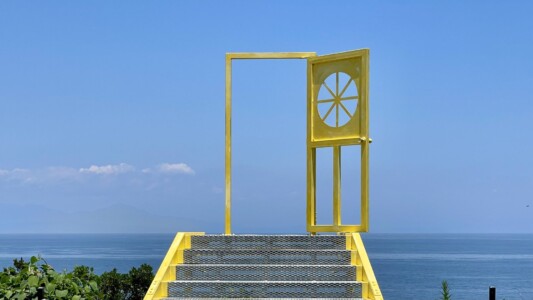 海を背景にした階段の上に立つ黄色いドアのオブジェ。ドアは半開きになっており、その向こうに広がる青い空と海が見える。階段の両側には緑の植物が茂り、遠くには雲が浮かぶ山々がぼんやりと見える。幻想的で非現実的な雰囲気を醸し出している。