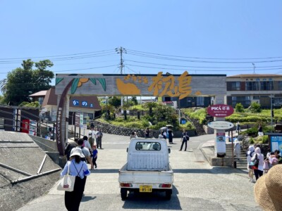 「ようこそ初島へ」と書かれた大きな看板が入口に掲げられた港の風景。看板にはオレンジのイラストが描かれている。入り口には観光客が行き交い、右側には「PICA初島」と書かれた案内板が見える。背景には建物が立ち並んでいる。手前には白い軽トラックが停まっており、日差しが強い中で多くの人々が観光を楽しんでいる様子がうかがえる。