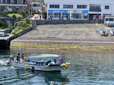 港の船着場に停泊している「DIVING CENTER」と書かれたダイビングボート。ボートには一人の男性が乗っており、エンジンを操作している。背景には階段やスロープがあり、建物の前には数名の人が立っている。
