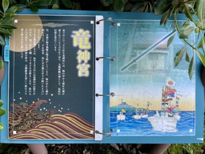 「竜神宮」と書かれた案内板がリングバインダーに綴じられている。左側のページには竜神宮に関する説明が白い文字で書かれ、背景には金色と赤色の波の模様が描かれている。右側のページには竜神宮のイラストがあり、手前に色とりどりの旗を掲げた船が海を進んでいる様子が描かれている。背景には神社の建物が見える。案内板は周囲の緑の葉に囲まれている。