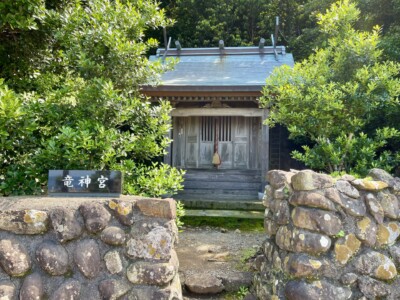 木々に囲まれた「竜神宮」の小さな神社。古い木製の建物には、正面に賽銭箱と鈴のついた縄が掛けられている。手前には「竜神宮」と書かれた黒い看板が石垣の上に設置されている。石垣は丸い石を積み上げて作られ、歴史を感じさせる。周囲の緑が生い茂る中、静かで神聖な雰囲気が漂っている。