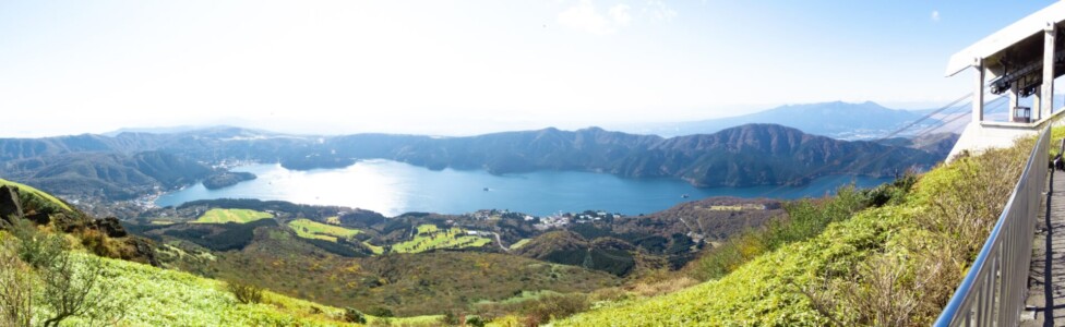 箱根駒ケ岳ロープウェーの頂上からのパノラマビュー。青々とした芦ノ湖が中央に広がり、その周囲には緑豊かな山々とゴルフ場が見える。湖の向こうには広がる山々と遠くの景色が一望でき、澄んだ空気と晴れた日の美しい景色が広がっている。右側には展望台の一部が見え、観光客がこの絶景を楽しんでいる様子が伺える。