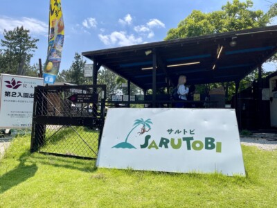 芝生の上に置かれた「サルトビ(SARUTOBI)」と書かれた看板が見えるエントランス。看板の隣には金網のゲートがあり、その奥に屋根付きの受付エリアが見える。受付には数名の人が並んでおり、背景には緑の木々と青空が広がっている。
