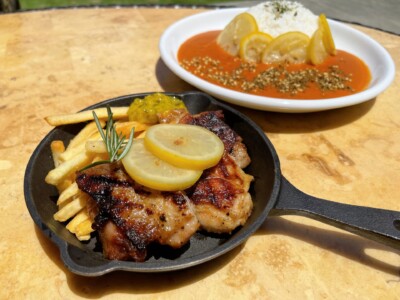 テーブルの上に並べられた美味しそうな料理。手前には鉄板に乗ったグリルチキンとフライドポテトがあり、チキンの上にはレモンスライスとローズマリーがトッピングされている。右奥にはレモンスライスが添えられたトマトソースカレーライスが白い皿に盛られている。どちらの料理も鮮やかで食欲をそそる。