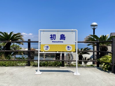 「初島 (Hatsushima)」と書かれた案内看板が立っている風景。看板の下部には黄色い背景に青い文字で「空 (Sky)」と「海 (Sea)」の矢印が描かれている。看板の背後には柵があり、その向こうにはヤシの木や海が広がっている。晴れた青空と美しい海の景色が見え、リゾート地らしい雰囲気が漂っている。