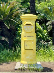 鮮やかな黄色の郵便ポストが緑の植物に囲まれて立っている。このポストには「郵便 (POST)」と「HATSUSHIMA」の文字が書かれている。周囲にはヤシの木やシダ植物が生い茂り、自然に囲まれた場所にあることがわかる。青空の下で、ポストの黄色が一際目立っており、観光地の一部として楽しむことができる。