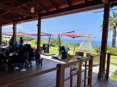 屋根付きのテラス席から海を望む景色。木製のテーブルとベンチに数人の人々が座り、くつろいでいる。外には赤いパラソルとバーベキューグリルが配置されており、緑の芝生とヤシの木が広がっている。遠くに海が見え、穏やかな雰囲気が漂う。