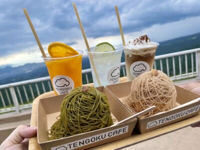 「TENGOKU CAFE」のドリンクとスイーツがトレイに乗せられている様子が写っています。トレイには、オレンジ色のドリンク、ライムのスライスが入った透明のドリンク、ホイップクリームがトッピングされたアイスコーヒーのカップが並んでいます。カップには「TENGOKU CAFE」のロゴが入っています。また、抹茶味と栗味のモンブランがそれぞれの紙容器に入っており、美しく盛り付けられています。背景には柵と山々が見え、外の景色を楽しみながらスイーツを楽しむことができるロケーションです。