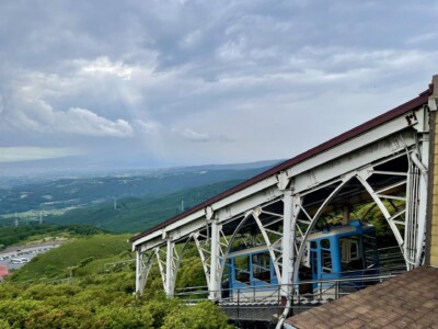 山の斜面に沿って設置されたケーブルカーの駅から、ケーブルカーが下りていく様子が写っています。駅の構造は金属製で、ケーブルカーは青と白のカラーリングです。背景には緑豊かな山々と広がる平野が見え、遠くには市街地や曇り空の下に差し込む光が見えます。手前には駐車場と赤い屋根の建物も確認できます。