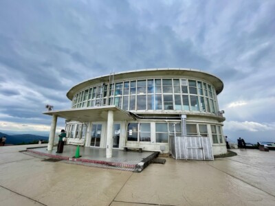 山頂展望デッキ「PANORAMA TERRACE 1059」・「TENGOKU CAFE」。建物はガラス張りで、内部が見えるようになっています。入り口には柱があり、建物の周囲には広いテラスがあります。テラスには観光客が数名おり、景色を楽しんでいる様子が伺えます。背景には山々と広がる景色が広がり、遠くには雲の間から光が差し込んでいます。