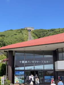 「十国峠パノラマケーブルカー」の駅舎が写っています。建物の入口には「十国峠パノラマケーブルカー」と書かれた大きな看板があり、多くの人々が入場しています。駅舎の上方にはケーブルカーのレールが山の斜面に沿って延びており、山頂には展望台が見えます。展望台は円形の建物で、ガラス張りになっており、周囲の緑豊かな景色と調和しています。