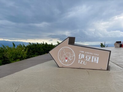伊豆国の矢印モニュメント。灰色の矢印型モニュメントが地面に設置されており、「十国峠 JUKKOKU TOGE 伊豆国 IZU」と書かれています。背景には青々とした植生と曇り空が広がっています。矢印の中には地図と位置を示す赤いポイントが描かれています。