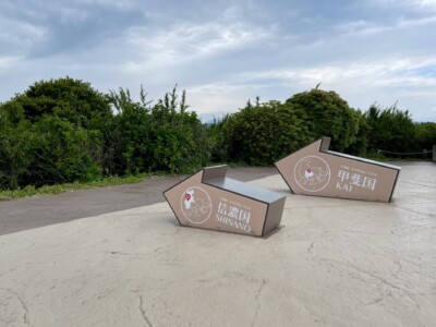 信濃国と甲斐国の矢印モニュメント。地面に設置された2つの灰色の矢印型モニュメントがあり、「十国峠 JUKKOKU TOGE 信濃国 SHINANO」と「十国峠 JUKKOKU TOGE 甲斐国 KAI」と書かれています。背景には緑豊かな植生と曇り空が広がっています。矢印の中には地図と位置を示す赤いポイントが描かれています。