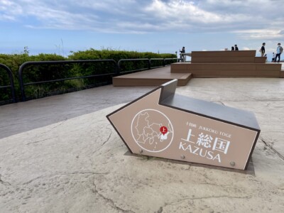 上総国の矢印モニュメント。灰色の矢印型モニュメントが地面に設置されており、「十国峠 JUKKOKU TOGE 上総国 KAZUSA」と書かれています。背景には緑豊かな植生と曇り空が広がり、観光客が散策しています。矢印の中には地図と位置を示す赤いポイントが描かれています。