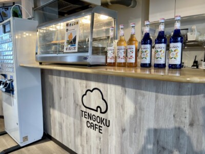 「TENGOKU CAFE」のカウンターの一部が写っています。カウンターの上には、焼き峠チュロスがディスプレイされた温蔵ショーケースがあり、その隣にはシロップボトルが並んでいます。カウンターの側面には「TENGOKU CAFE」のロゴが描かれており、明るい木目調のデザインが施されています。左側には券売機が見えます。全体的に清潔で整然とした雰囲気です。